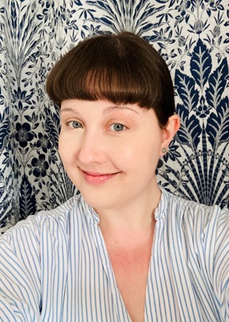 Photo of Lydia Savory, brunette with fringe wearing striped shirt and standing against patterned background