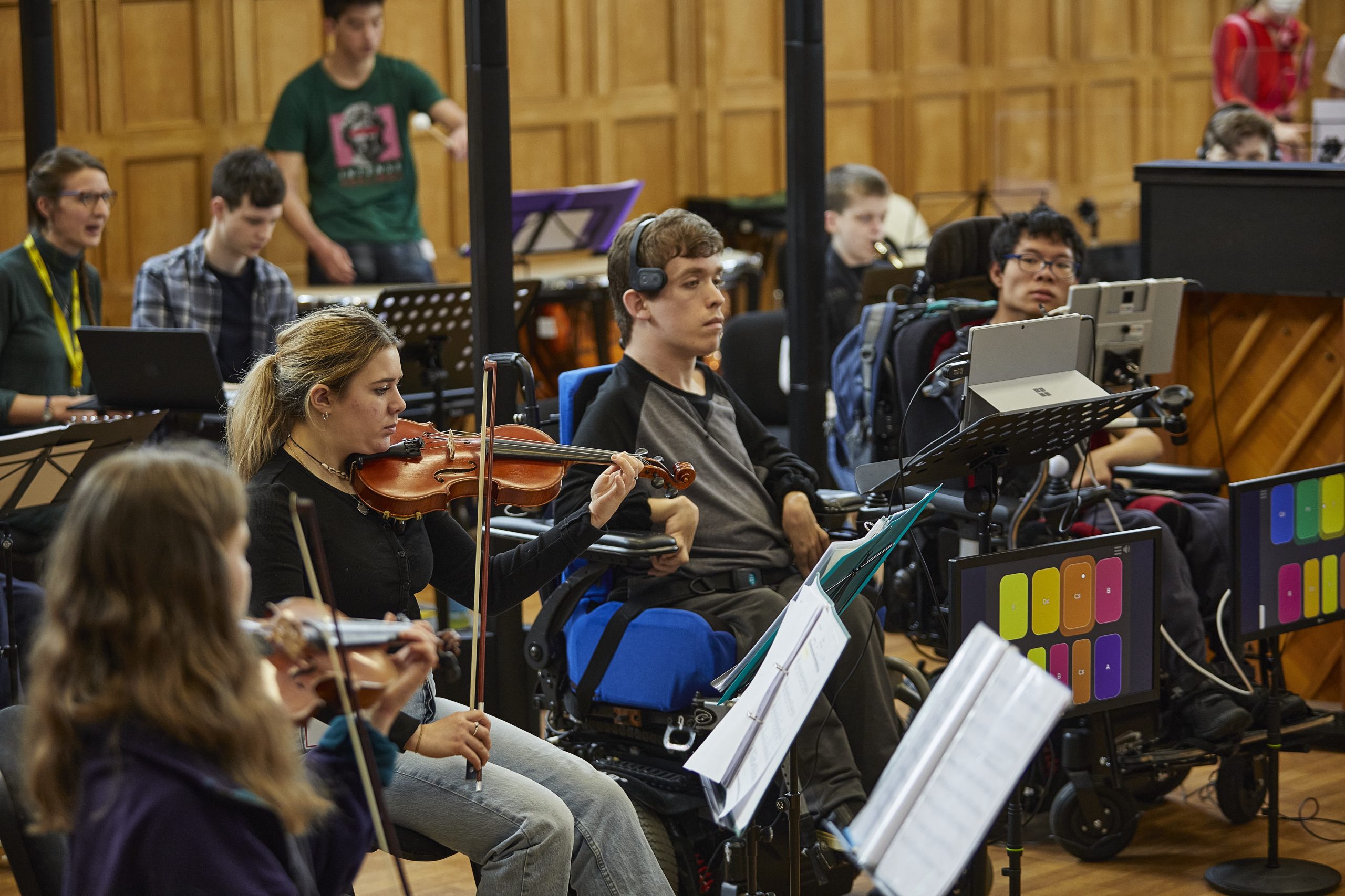 Members of the National Open Youth Orchestra rehearse together in a large room. Some musicians are playing traditional instruments like violins, while others use adaptive instruments such as the Clarion mounted on wheelchairs. The inclusive ensemble features both disabled and non-disabled musicians.