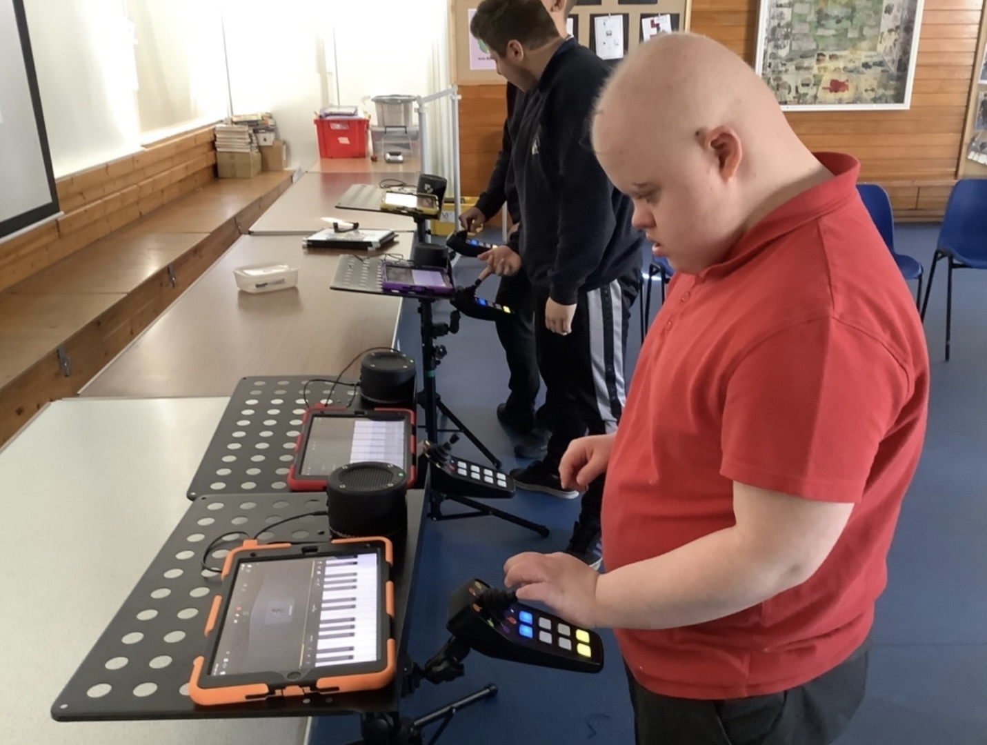 A young person in a red polo shirt is using a CMPSR, an accessible digital musical instrument, in a classroom setting. The CMPSR is connected to a tablet showing a digital keyboard interface. Another person in the background is also using similar equipment.